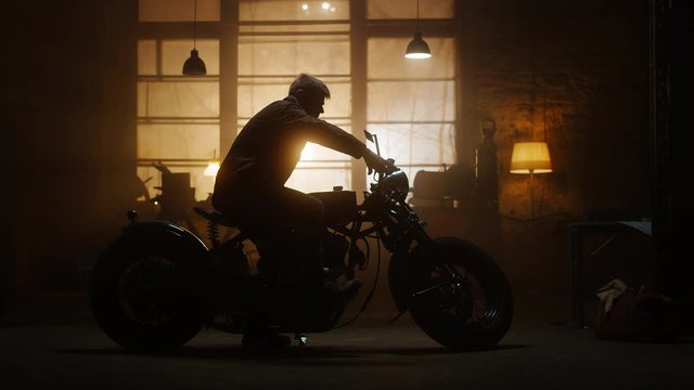 Custom Bobber Motorbike Standing in an Authentic Creative Workshop. Rider Gets on a Bike. Vintage Style Motorcycle Under Warm Lamp Light in a Garage.