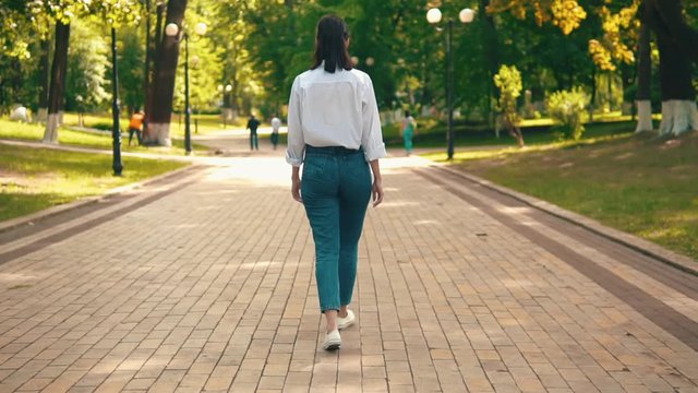 unrecognizable female in casual white shirt and sneakers walking in city park summer season