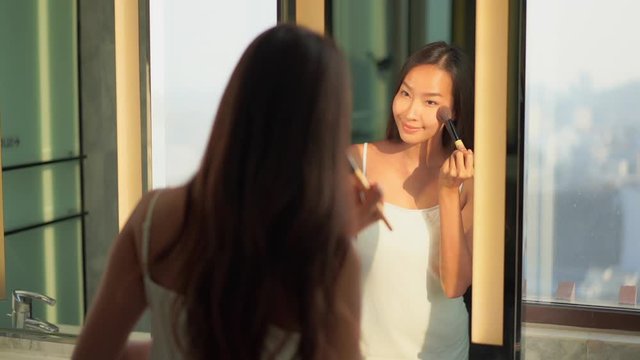 An attractive young Asian woman while looking in a  mirror applies the finishing touches to her makeup with a complexion brush.