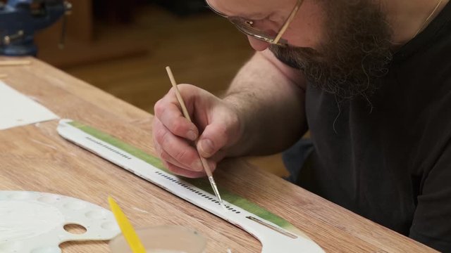 bearded Sunday school teacher paints vintage jetliner model fuselage in white on wooden table with palette slow motion closeup