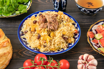 National Uzbek pilaf with meat, achichuk salad of tomato, cucumber, onion in plate with traditional pattern, cilantro, cherry tomatoes, garlicbread tortilla - patir on dark wooden table Top view