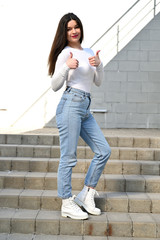 Photo of a caucasian brunette girl in a white blouse and blue jeans stands on the stairs with a smile against the background of a gray wall of a building on a sunny spring day.