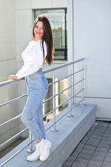 Fototapeta na wymiar Photo of a caucasian brunette girl in a white blouse and blue jeans stands near the railing with a smile against the background of a gray wall of a building on a sunny spring day.