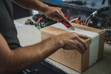 Hands of a man seen opening a brown cardboard box using a red utility knife. Service bench in the...