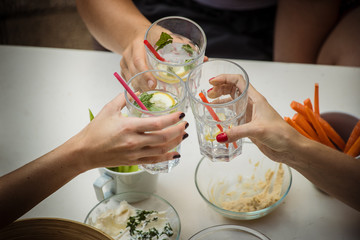 Hands of three women viewed from above seen cheering or toasting with glasses of gin tonic drink. Other healthy foods are seen around, like dips and carrots