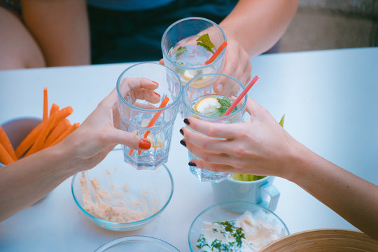 Hands Of Three Women Viewed From Above Seen Cheering Or Toasting With Glasses Of Gin Tonic Drink. Other Healthy Foods Are Seen Around, Like Dips And Carrots