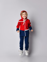 Full growth portrait of active happy laughing blond kid boy in blue and red sportswear with his hood on his head jumps