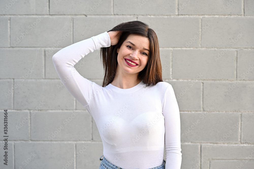 Wall mural A caucasian portrait of a pretty brunette girl in a white blouse stands near the wall of a building on a spring day in the city. Model with pleasure and smile posing.