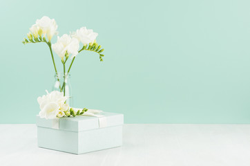 Romantic holiday background with spring flowers freesia in glass vase and closed gift box with ribbon in green mint menthe interior on white wood board.