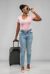 travel, tourism and vacation concept - happy african american young woman in sunglasses with carry-on bag over grey background