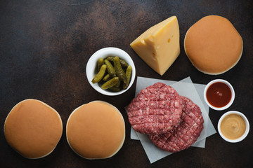 Raw marbled beef cutlets, burger buns, slab of cheese and pickles. Flatlay on a dark brown metal background, horizontal shot with space