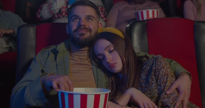 Front view of pretty girl sleeping on her boyfriend shoulder during movie. Bearded guy eating popcorn and hugging girlfriend, looking at her and smiling at cinema.