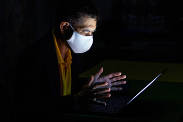 An Asian old business man uses his computer notebook while wearing face mask to connect with...