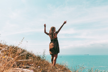 Beautiful slender tattooed woman with blonde dreadlocks in a dress rejoices on a hill. Sea View...