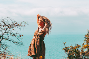 Beautiful slender tattooed woman with blonde dreadlocks in a dress rejoices on a hill. Sea View Travel and Vacation lifestyle Beautiful landscape