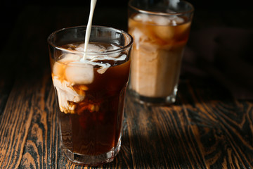 Pouring of milk into cold coffee in glass on wooden background