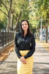 portrait of a young elegant business woman standing on the sidewalk among the trees in a black shirt and yellow skirt