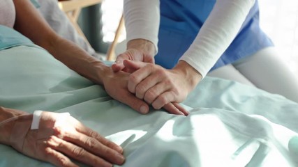 Unrecognizable friendly woman doctor sitting by patient in bed in hospital, holding hands. - Powered by Adobe