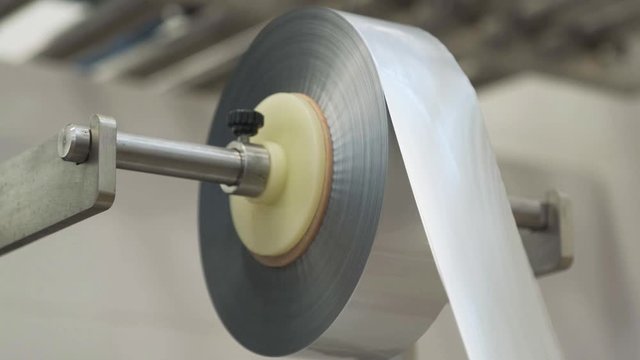 A roll of shiny silver aluminum foil rotates on a metal stick, unwinding the required amount. Material for the manufacture of portioned bags. Roll of mica rotation. Manufacturing process.