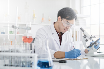 A male scientist with black hair wearing white coat and protective glassware writing and looking into a microscope in a laboratory setting with test tubes. 
