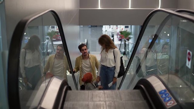 Young attractive caucasian family with suitcase going up by the escalator with their pretty daughter hold by mothers hand at the airport. Happy family vacation together