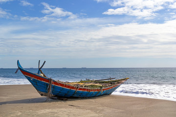 Naklejka na ściany i meble Wooden fishing boat on a deserted beach on a background of foam waves and a blue sky with clouds on a sunny day. Copy space