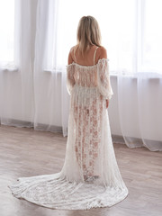 Young pretty blond woman bride, wearing long white lace bridal peignoir, standing in the light room with back to camera, looking at the window, on the wedding morning