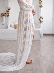 Close-up picture of the bottom of beautiful white lace peignoir night gown on young woman, standing in the middle of light bedroom with white furniture.