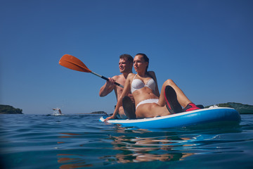 Young and beautiful couple of lovers sitting on a surfboard in the open ocean on a background of sky.