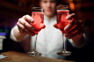 bartender in a white shirt holds out two glasses with red champagne or cocktail against the background of the bar