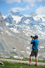 Side view of tourist using camera to take photo of beautiful mountains scenery on background. Mountain hiking. Sport tourism in Alps. Concept of travelling and professional photography.