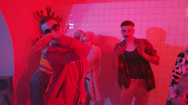 Young eccentric man in trendy wear and sunglasses dancing expressive vogue with group of stylish friends in room with color neon light and fog