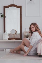 young beautiful woman in white shirt sitting on the floor near the bed and looking to the camera