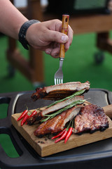 Fried pork ribs with a fried crust with spices, rosemary and chili pepper on a wooden board on the outdoor  with fork and hand. Meat on grill. Background image, copy space