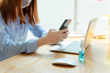 Asian woman wearing mask staying at home playing smartphone relaxing emotion, Prevent the spread of coronavirus quarantine work desk with computer and protective surgical mask.