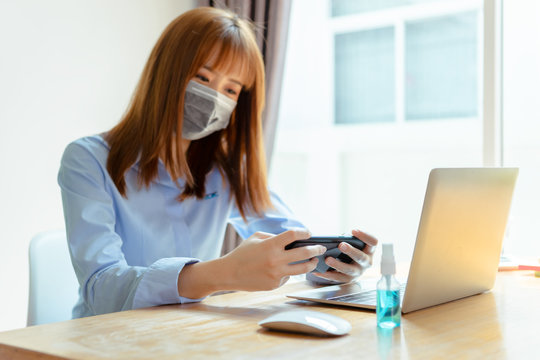 Asian woman wearing mask staying at home playing smartphone relaxing emotion, Prevent the spread of coronavirus quarantine work desk with computer and protective surgical mask. - Powered by Adobe