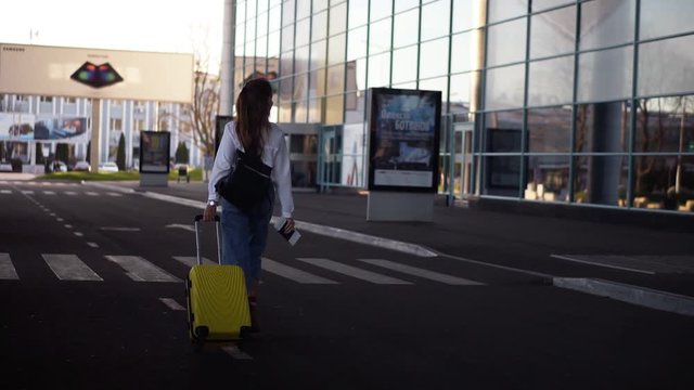 Stylish woman walk with trolley yellow case by empty airport terminal outside road, slender female wearing jeans and brown boots. Back side view