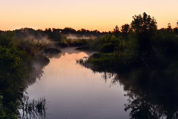 Obraz na płótnie Canvas River in the early morning at dawn. Delicate dawn sky and fog rising above the water, lush greenery on the banks. Summer spring wild landscape by the river. Selective soft focus.