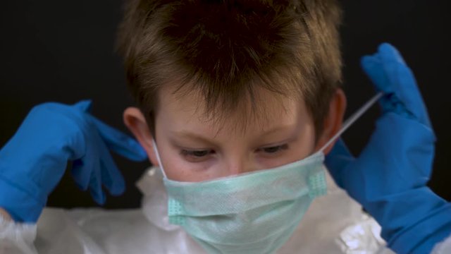 Beautiful cute blonde child in a white protective medical outfit in blue gloves put mask, respirator on face, glasses, hood close up on black background. Quarantine by reason of coronavirus concept.