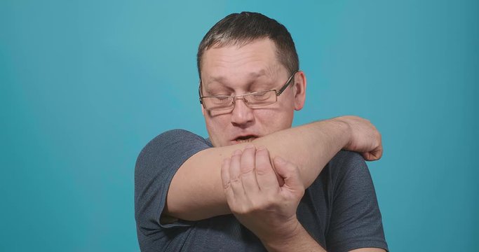 man with glasses in grey t-shirt sneezes into arm bend standing against blue background slow motion closeup