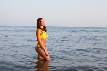 Beautiful tanned woman in yellow swimsuit stands in the sea bathing on the beach