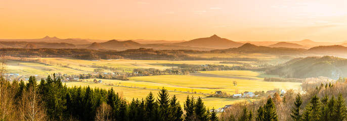 Fototapeta premium Hilly landscape illuminated by evening sunset. Green grass fields and hills on the horizont. Vivid spring rural countryside. Ralsko Mountain, Czech Republic
