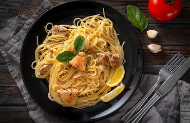 pasta, spaghetti with chicken fillet, lemon slices, herbs, tomato, garlic, on a black plate on a dark wooden background,  fork, knife, napkin,  top view