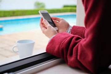 A woman reading the news on her phone while drinking coffee and wearing pajamas and a robe by the window