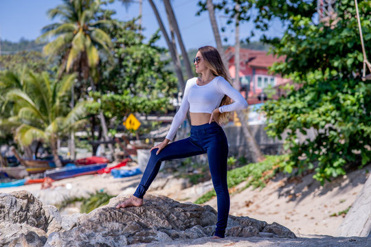 Fashion picture in Thailand with female model, dark blue jeans, white top, and sunglasses, summer time, background palms and beach.