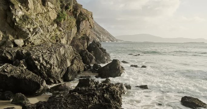 Pictorial rocky cliffs in earthy brown and washed out black are hit by waves rolling in and by foamy spray. Cliffs of Achill Island.
