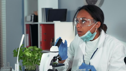 Multiracial research scientist getting shocked with the result of the experiment seen under the microscope. Woman is in safety glasses and lab coat. Coronavirus pandemic concept.