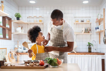 Happy family with African father and son dress up together before cooking in the white kitchen. Single Dad Chef with black kid helper in yellow shirt preparing food and looking at Each Other at home. 