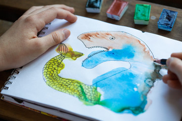 Child's drawing of watercolor paints in a sketch notebook, children's creativity concept. Child's hands next to the picture. Monotype. Drawing of an unusual fairytale animal.