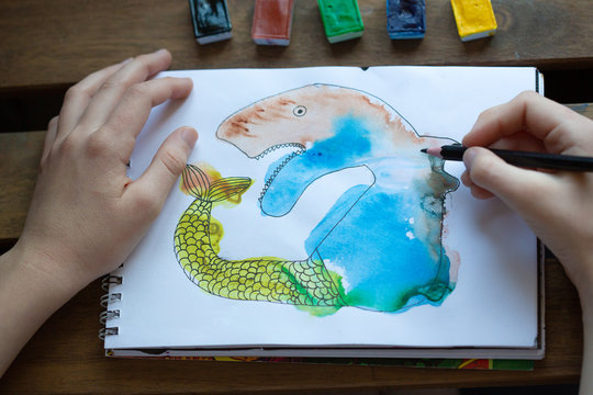 Child's drawing of watercolor paints in a sketch notebook, children's creativity concept. Child's hands next to the picture. Monotype. Drawing of an unusual fairytale animal.